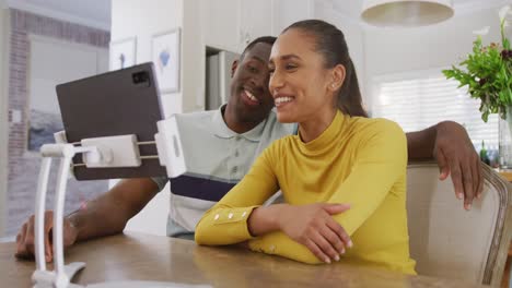 Video-of-happy-diverse-couple-watching-tablet-together-and-laughing-sitting-in-kitchen
