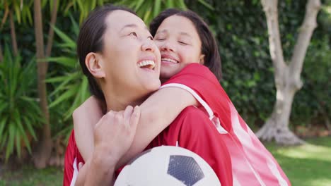 Video-Einer-Glücklichen-Asiatischen-Mutter-Und-Tochter-In-Fußballtrikots,-Die-Sich-Im-Garten-Umarmen