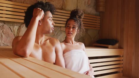 Video-of-relaxed-diverse-couple-wearing-towels-sitting-and-talking-in-sauna-room-at-health-spa