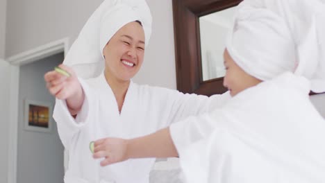 Video-Einer-Glücklichen-Asiatischen-Mutter-Und-Tochter-In-Roben,-Die-Eine-Gurkenmaske-Machen-Und-Spaß-Haben