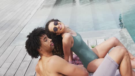Video-of-happy-diverse-couple-relaxing-and-talking-sitting-at-the-poolside-of-holiday-home