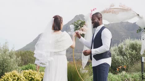 Video-of-happy-diverse-bride-and-groom-holding-hands,-smiling-and-dancing-at-outdoor-wedding