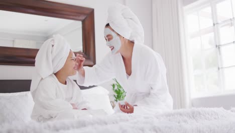 Video-Einer-Glücklichen-Asiatischen-Mutter-Und-Tochter-In-Bademänteln,-Die-Ihr-Gesicht-Mit-Feuchtigkeit-Versorgen-Und-Spaß-Haben