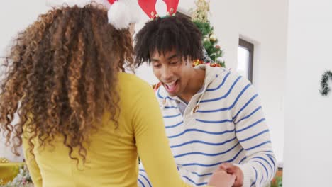 Video-of-happy-diverse-couple-in-reindeer-antlers-and-santa-hat-dancing-by-christmas-tree-at-home