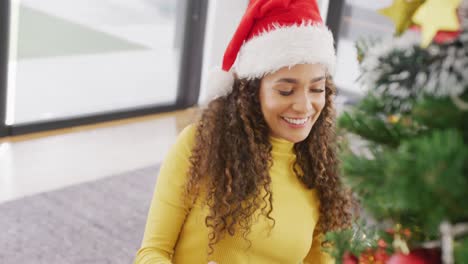 Video-of-happy-biracial-woman-in-santa-hat-smiling-and-decorating-christmas-tree-at-home