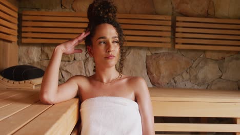 Video-of-relaxed-biracial-woman-wearing-towel-sitting-in-sauna-room-at-health-spa