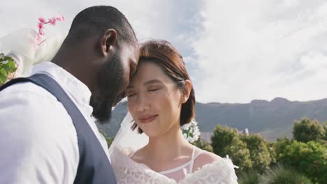 Video-of-happy-diverse-bride-and-groom-touching-heads-together-and-smiling-at-outdoor-wedding