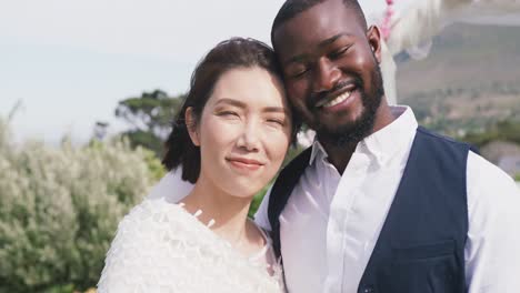 Video-portrait-of-happy-diverse-bride-and-groom-embracing-and-smiling-to-camera-at-outdoor-wedding