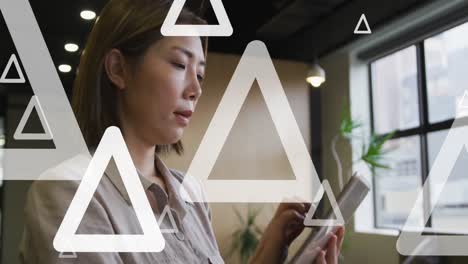 Animación-De-Triángulos-Blancos-Moviéndose-Sobre-Una-Empresaria-Asiática-Usando-Una-Tableta-Digital-En-La-Oficina