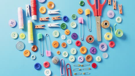 Video-of-scissors,-buttons,-pins,-clips,-pegs-and-cotton-reels-in-a-heart-shape-on-blue-background