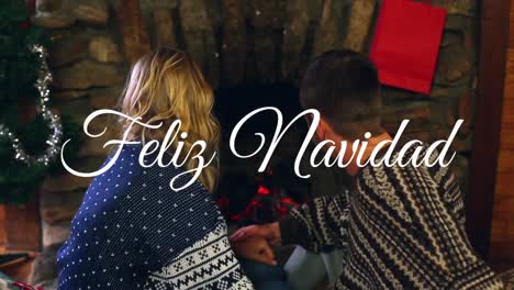 Animation-of-feliz-navidad-christmas-greetings-text-over-caucasian-couple-by-fireplace-at-christmas