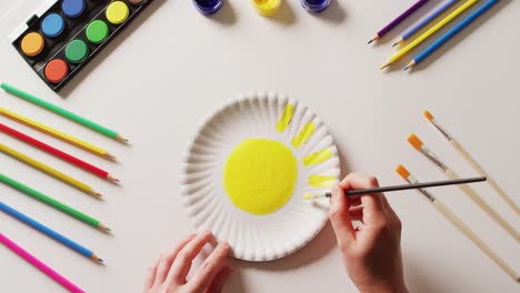 Video-of-hands-painting-design-on-paper-plate,-with-art-materials-arranged-on-white-background
