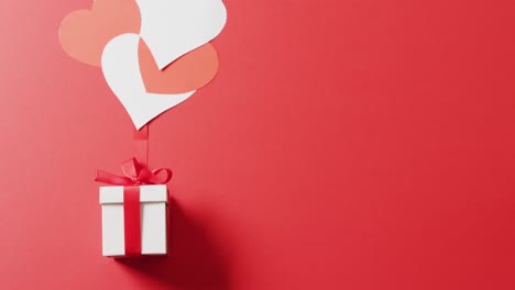 Video-of-white-gift-box-with-red-ribbon-and-red-and-white-hearts,-on-red-background-with-copy-space