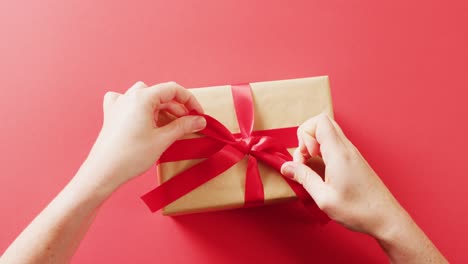 Video-of-hands-tying-red-ribbon-on-gift-box-wrapped-in-brown-paper,-on-red-background