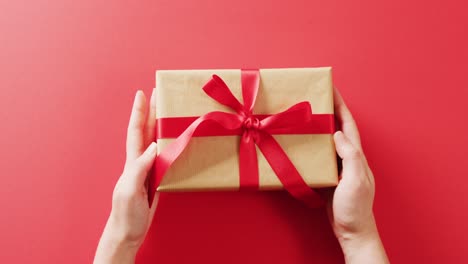 Video-of-hands-placing-gift-box-wrapped-in-brown-paper-and-red-ribbon-on-red-background