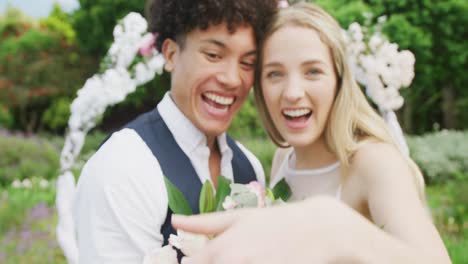 Portrait-of-happy-diverse-couple-holding-hands-with-ring-on-sunny-day-at-wedding