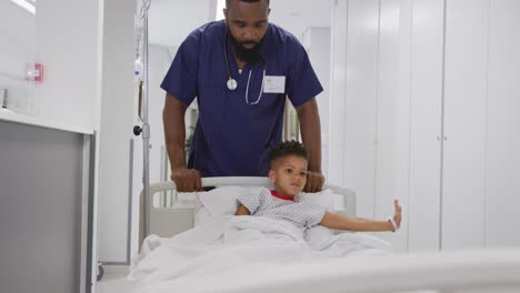 Médico-Varón-Afroamericano-Caminando-Con-Un-Paciente-Infantil-Acostado-En-La-Cama-En-El-Hospital