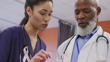 Two-diverse-male-and-female-doctors-wearing-cancer-awareness-ribbons-talking-at-hospital
