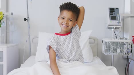 Portrait-of-happy-african-american-boy-patient-sitting-up-in-bed-at-hospital-smiling