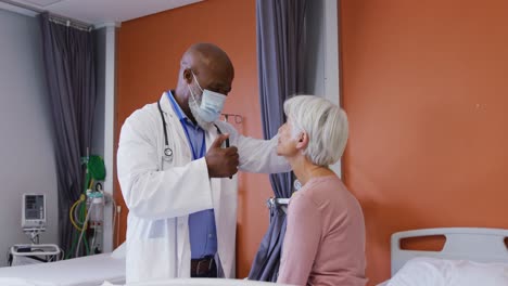 Médico-Varón-Afroamericano-Usando-Una-Linterna-Examinando-Los-Ojos-De-Una-Paciente-Caucásica-Mayor