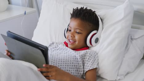 Glücklicher-Afroamerikanischer-Junge-Mit-Tablet-Und-Kopfhörern-Im-Bett-Im-Krankenhaus