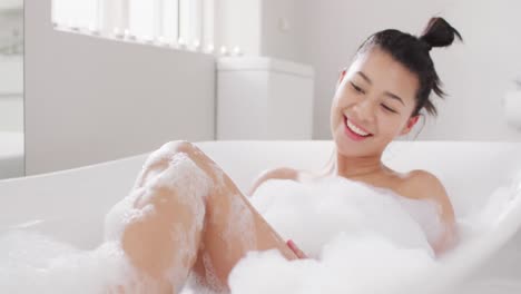 Video-of-portrait-of-smiling-biracial-woman-sitting-in-bathtub-in-bubble-bath-in-bathroom