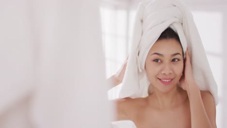 Video-of-portrait-of-smiling-biracial-woman-with-towel-on-hair-looking-in-mirror-in-bathroom