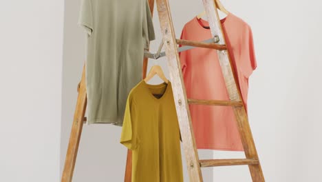 Video-of-close-up-of-mustard,-green-and-orange-t-shirts-hanging-from-ladder-on-white-background