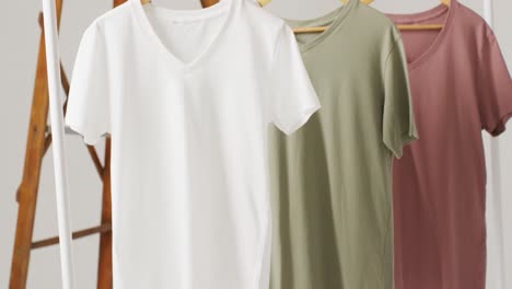 Video-of-close-up-of-white,-beige-and-gren-t-shirts-hanging-on-white-background