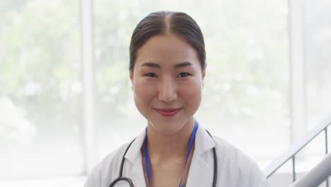 Video-portrait-of-happy-asian-female-doctor-smiling-in-hospital-corridor
