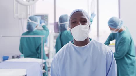 Video-portrait-of-african-american-male-surgeon-in-mask-smiling-in-operating-theatre,-copy-space