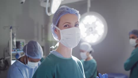 Video-portrait-of-caucasian-female-surgeon-in-face-mask-smiling-in-operating-theatre,-copy-space