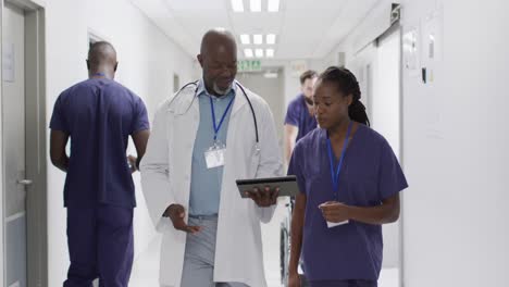 Video-of-african-american-male-and-female-doctor-walking-in-hospital-corridor,-looking-at-tablet