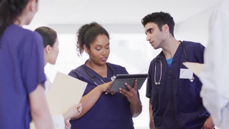 Video-of-diverse-group-of-male-and-female-doctors-looking-at-tablet-talking-in-hospital-corridor