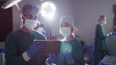 Video-of-diverse-male-and-female-surgeon-in-discussion,-looking-at-tablet-in-operating-theatre