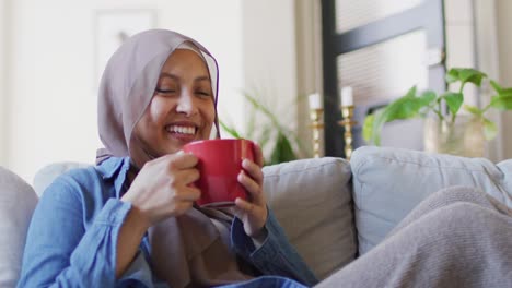 Video-of-smiling-biracial-woman-in-hijab-drinking-coffee-relaxing-on-sofa-in-living-room-at-home