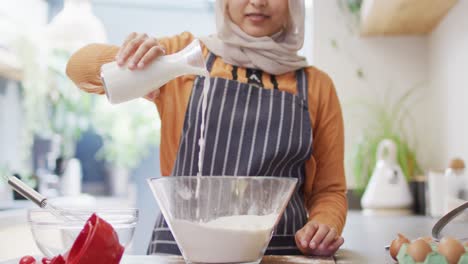 Video-of-happy-biracial-woman-in-hijab-baking-in-kitchen-at-home,-pouring-milk-into-mixing-bowl