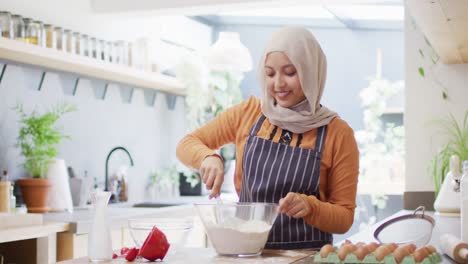 Video-De-Una-Mujer-Birracial-Feliz-Con-Hijab-Horneando-En-La-Cocina-De-Casa,-Mezclando-Masa-De-Pastel-En-Un-Tazón