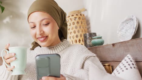 Video-of-happy-biracial-woman-in-hijab-drinking-coffee-and-using-smartphone-at-home
