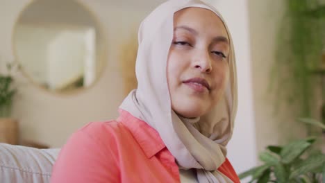 Video-Einer-Nachdenklichen-Gemischtrassigen-Frau-Im-Hijab,-Die-Zu-Hause-Im-Wohnzimmer-Schreibt-Und-Lächelnd-Wegschaut
