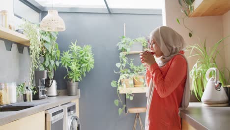 Video-Einer-Glücklichen-Gemischtrassigen-Frau-Im-Hijab,-Die-Zu-Hause-In-Der-Küche-Tee-Trinkt
