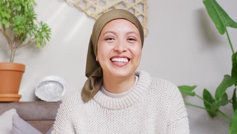 Video-of-happy-biracial-woman-in-hijab-making-video-call-waving-to-camera-at-home