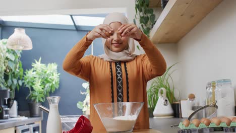 Video-De-Una-Mujer-Birracial-Sonriente-Con-Hijab-Horneando-En-La-Cocina-De-Su-Casa,-Rompiendo-Un-Huevo-En-Un-Tazón