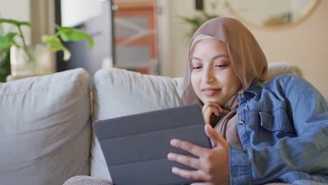 Video-of-happy-biracial-woman-in-hijab-using-tablet-relaxing-on-sofa-in-living-room-at-home