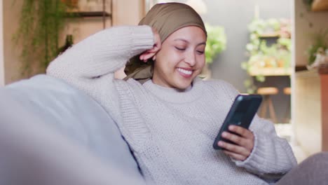 Video-of-happy-biracial-woman-in-hijab-using-smartphone-sitting-in-living-room-at-home