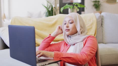 Video-of-concentrating-biracial-woman-in-hijab-at-home-sitting-on-floor-working-on-laptop