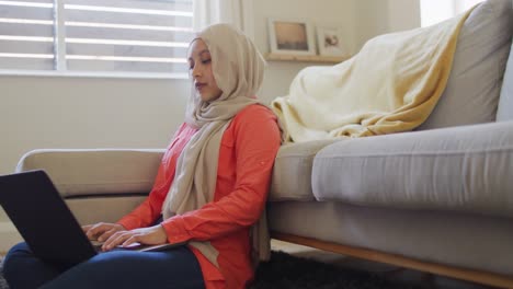 Video-De-Una-Mujer-Birracial-Concentrada-Con-Hijab-En-Casa-Sentada-En-El-Suelo-Trabajando-En-Una-Computadora-Portátil
