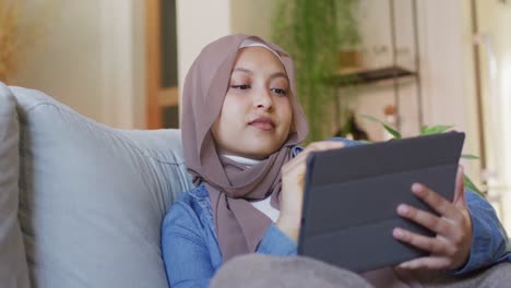 Video-of-happy-biracial-woman-in-hijab-using-tablet-relaxing-on-sofa-in-living-room-at-home