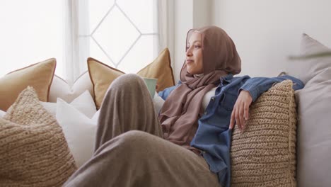 Video-Einer-Lächelnden-Gemischtrassigen-Frau-Im-Hijab,-Die-Kaffee-Trinkt-Und-Sich-Zu-Hause-Auf-Dem-Sofa-Entspannt