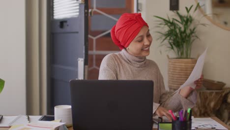 Video-of-smiling-biracial-woman-in-hijab-at-home-sat-at-desk-looking-at-paperwork-and-using-laptop